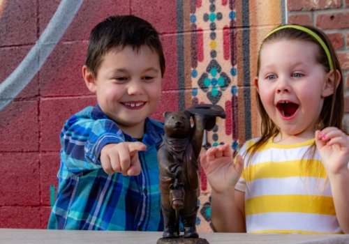 Two excited children in front of a small bear figurine.