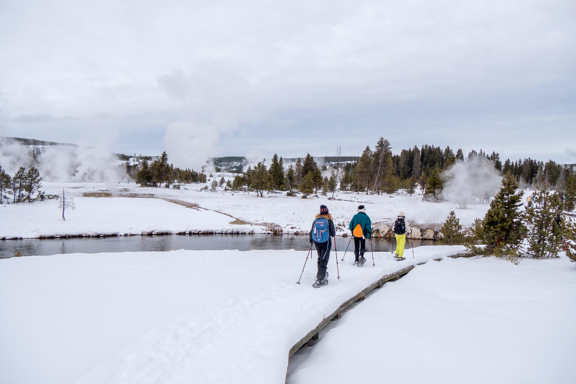 Three people snowshoeing on the boardwalks in the Upper Geyser Basin near the Firehole River.