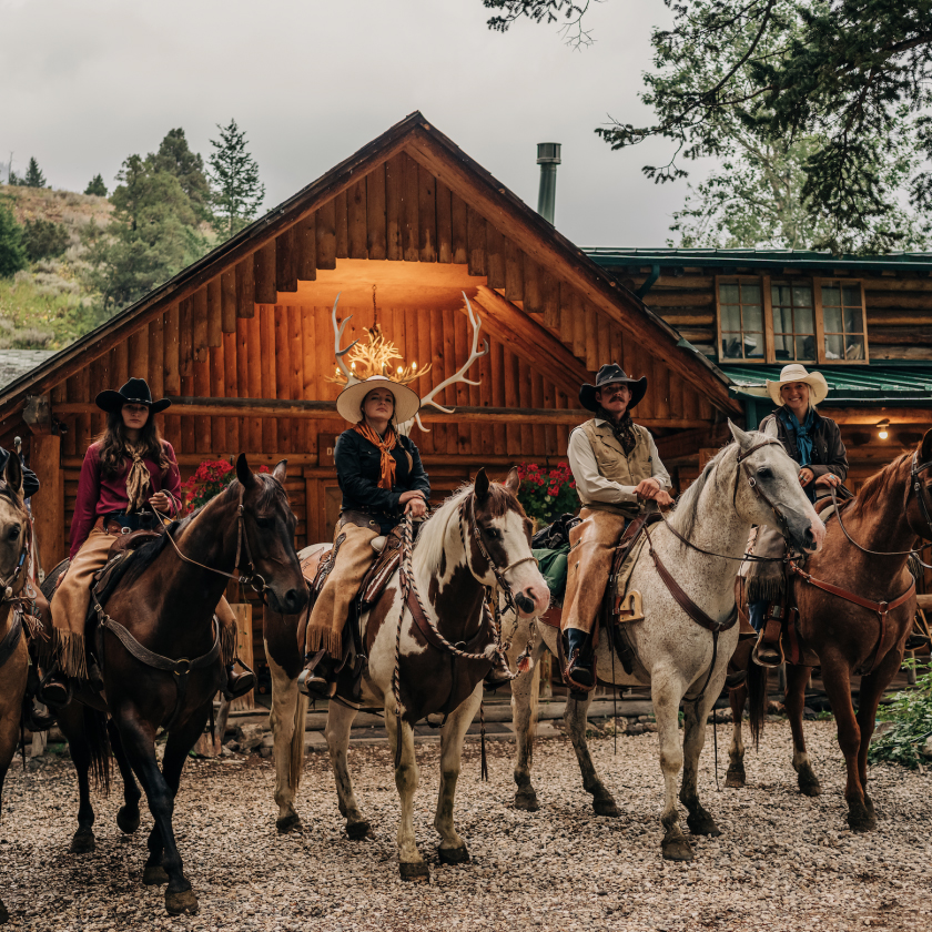 A cheerful group of horse riders poses in front of a charming log cabin, surrounded by trees and open space.