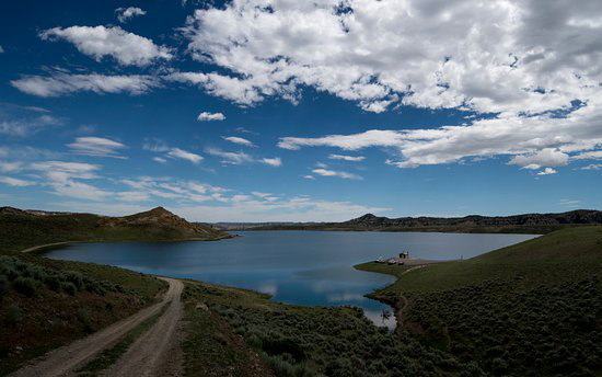 A rustic dirt road meanders toward a peaceful lake, bordered by lush hills all around.