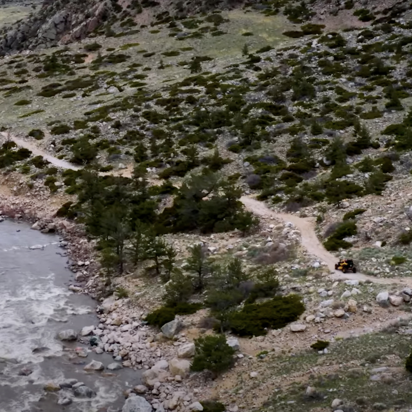 A beautiful mountain road curves beside a sparkling river, with majestic mountains rising in the distance.