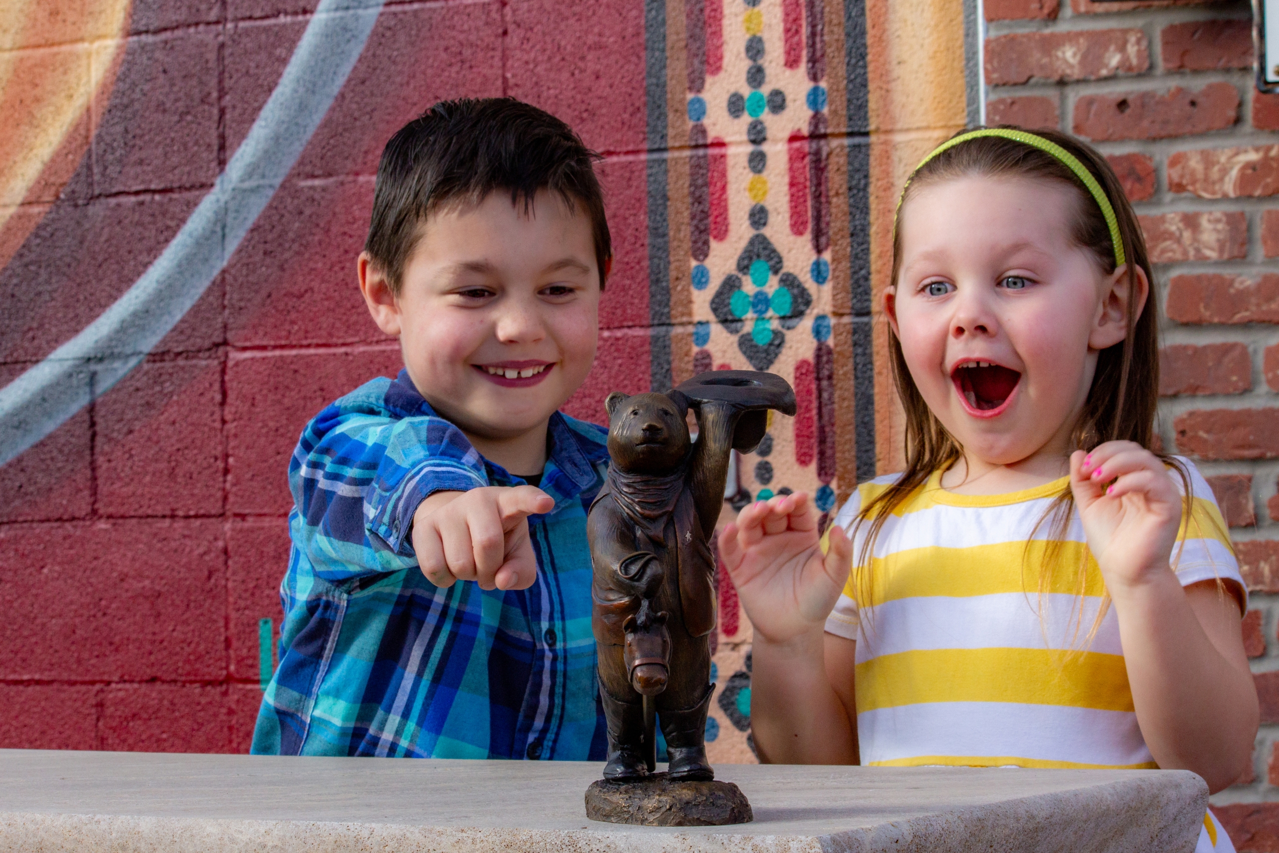 Two children excited while looking at a tiny copper statue of a bear with a hat and a old fashioned light.