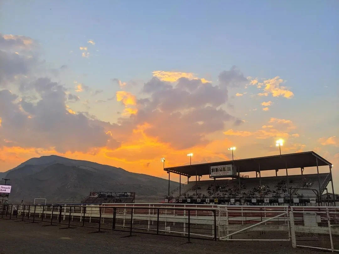 A vibrant sunset casts warm hues over the rodeo stadium, creating a stunning backdrop for the evening events