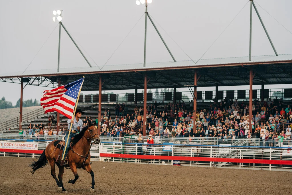 A cowboy on a horse holds an American flag high as he rides before a lively crowd