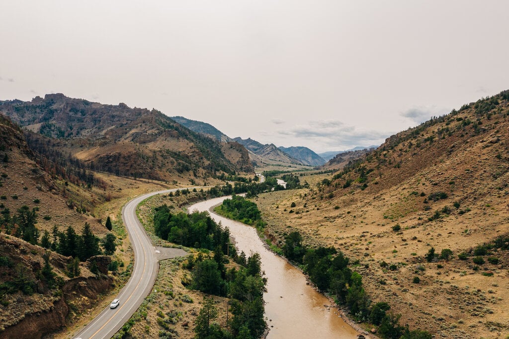 A winding road snakes through towering mountains, with a river flowing gracefully beside it, creating a picturesque view.